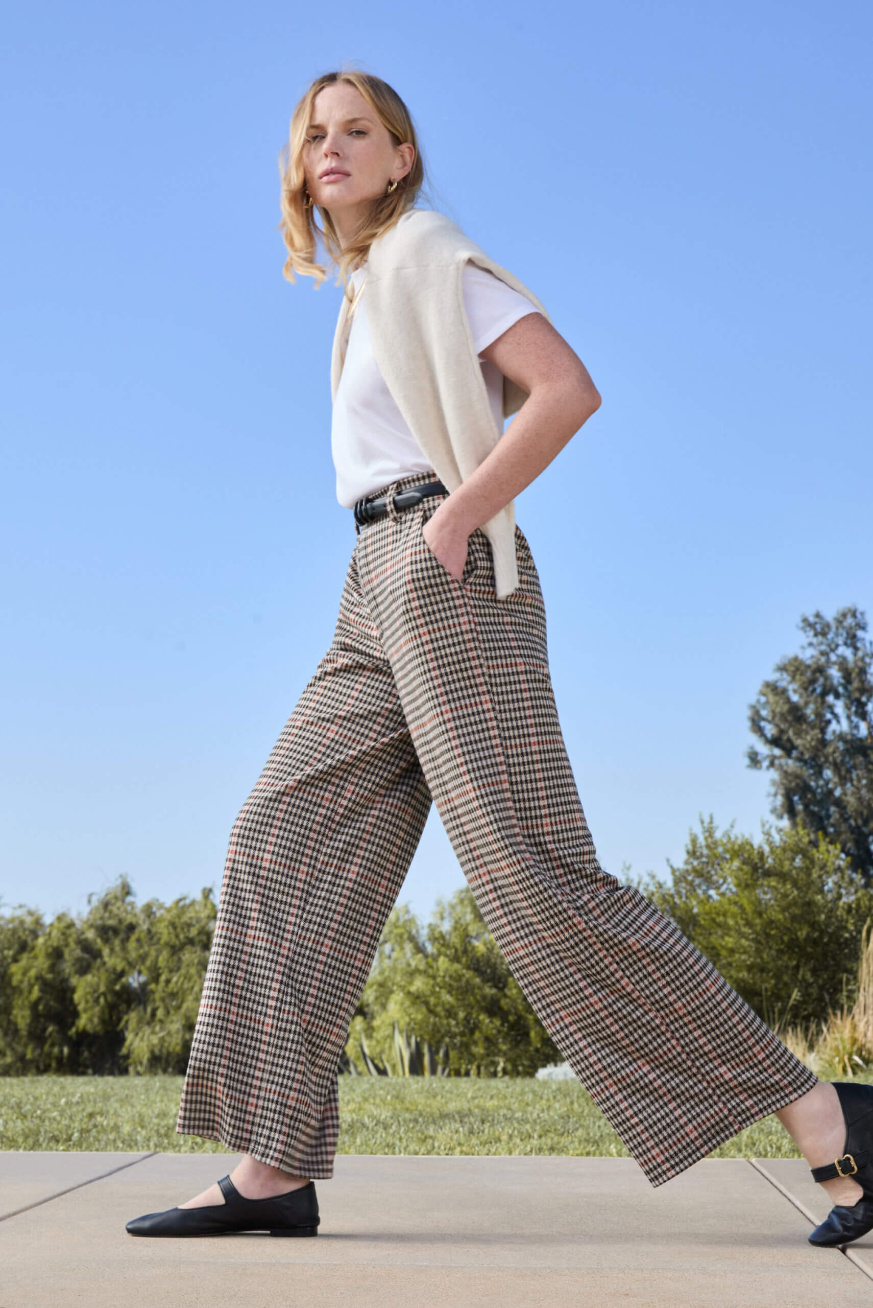A woman wearing drape-fit houndstooth pants, a white polo with a sweater over her shoulders, and black Mary Janes.