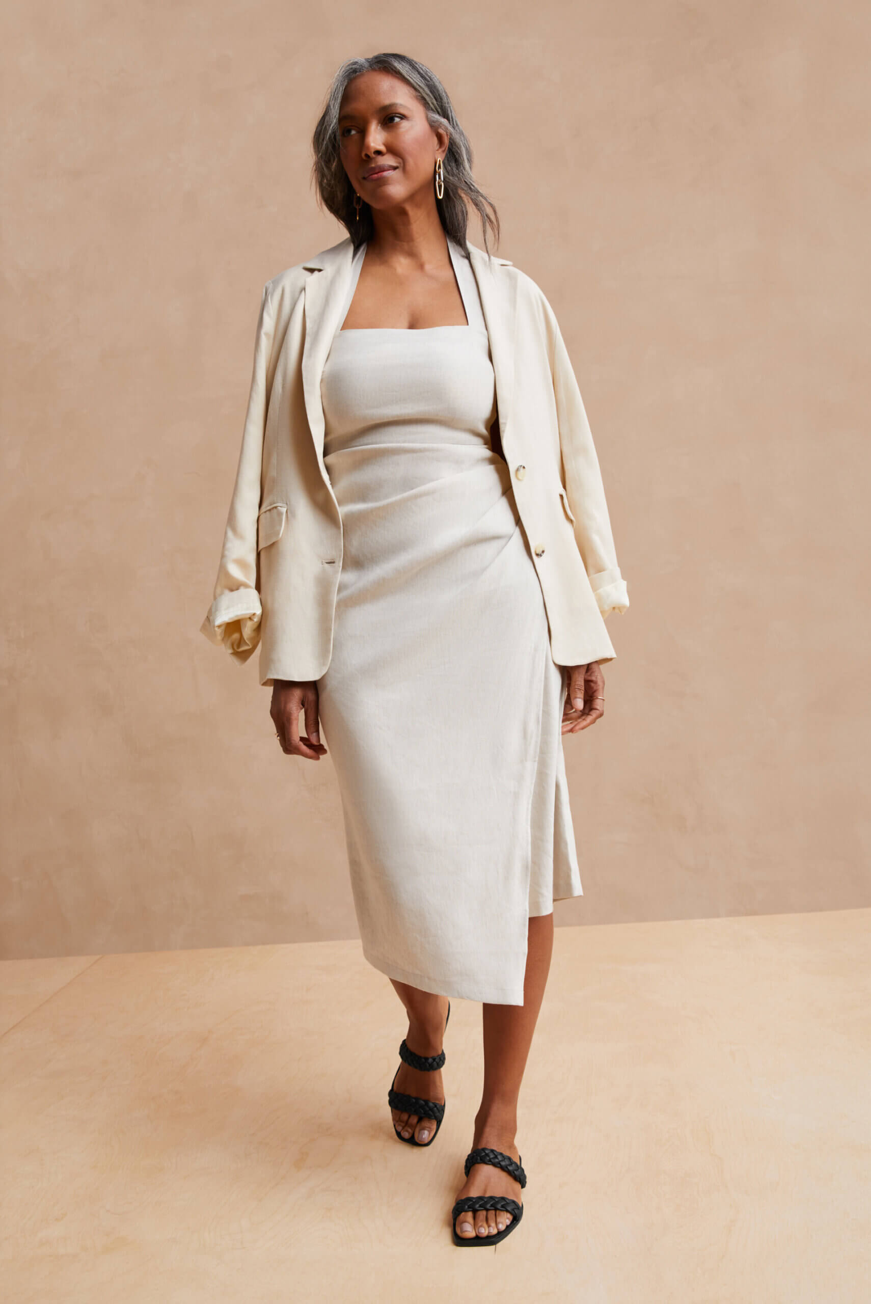 Black woman standing and wearing long white dress, cream blazer and black sandals.  