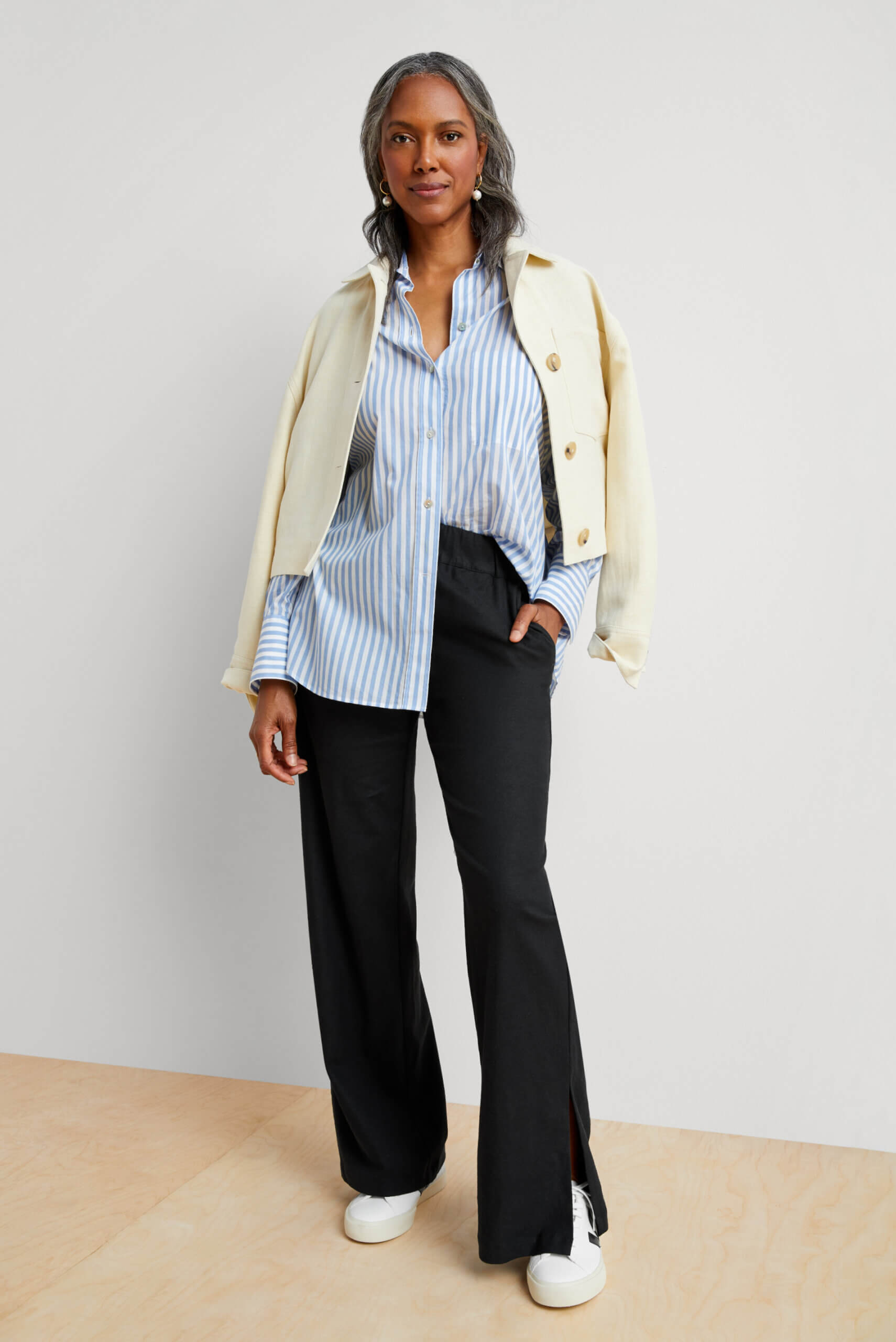 Black woman in striped, blue button-down shirt half tucked into dark blue pants with a cream blazer resting on her shoulders. 
