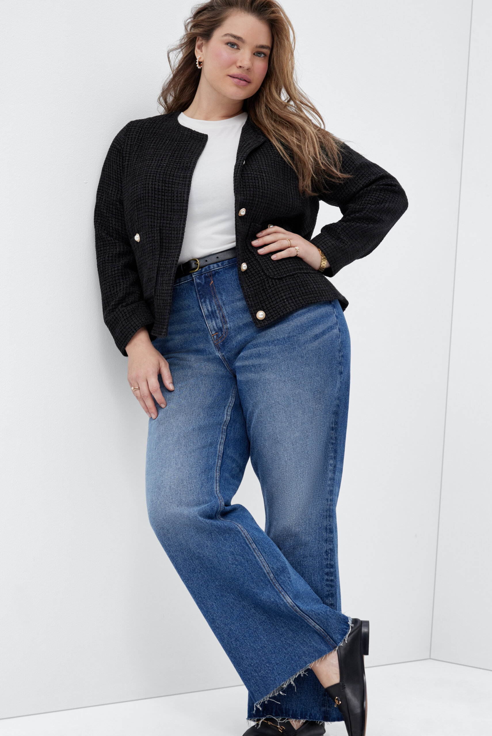 A plus-size woman wears an open black blazer over a white t-shirt and blue flare jeans.