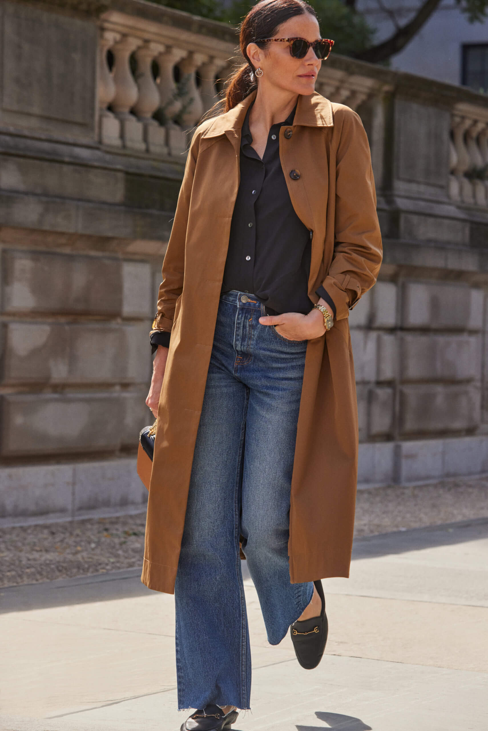Alt text: A woman walks wearing sunglasses, a black button-down shirt and blue flare jeans underneath a long brown coat. 